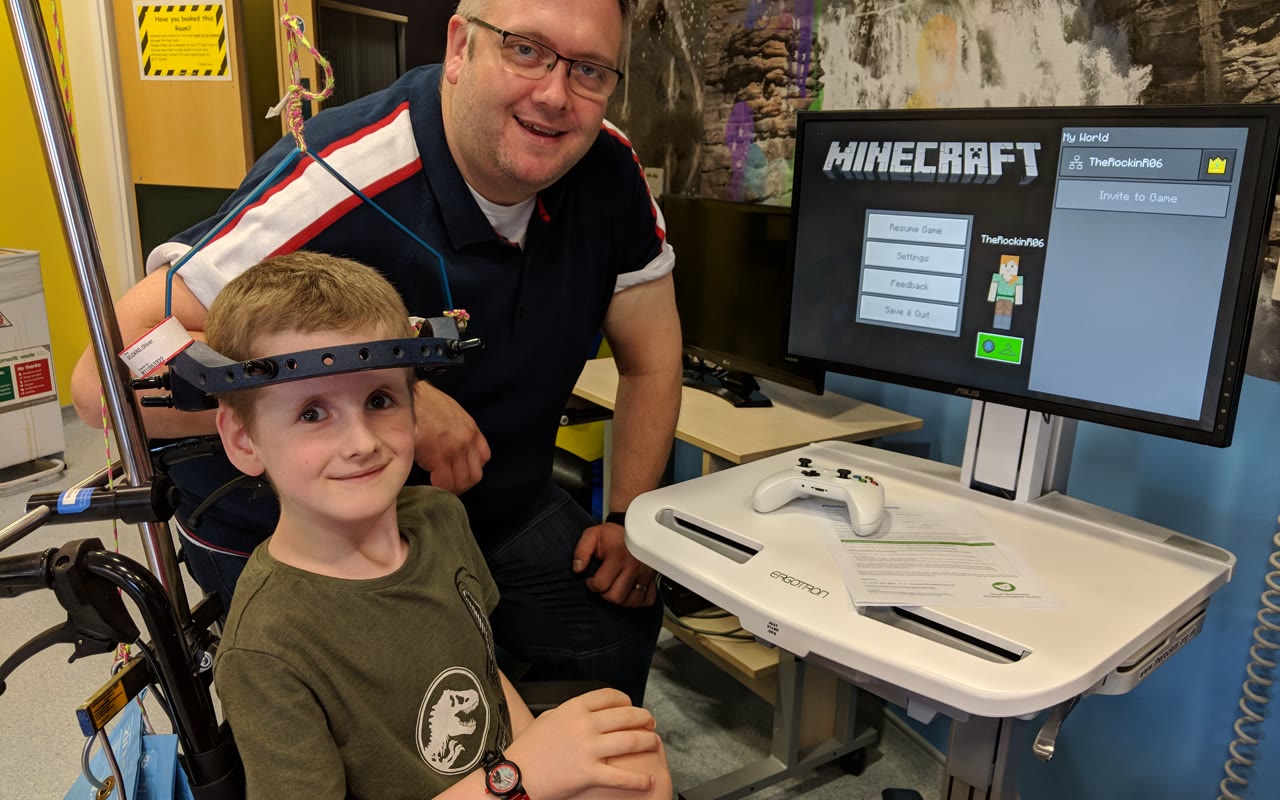 A boy in a head-frame and his dad smiling beside a Minecraft gaming cart in a hospital room.