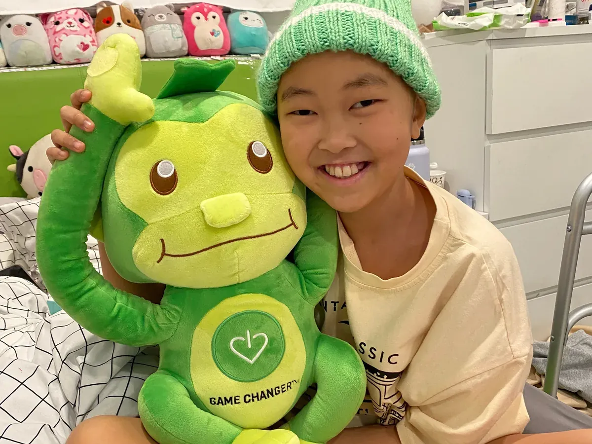 A young patient in a green beanie beaming as she hugs Hope, GameChanger's green monkey.